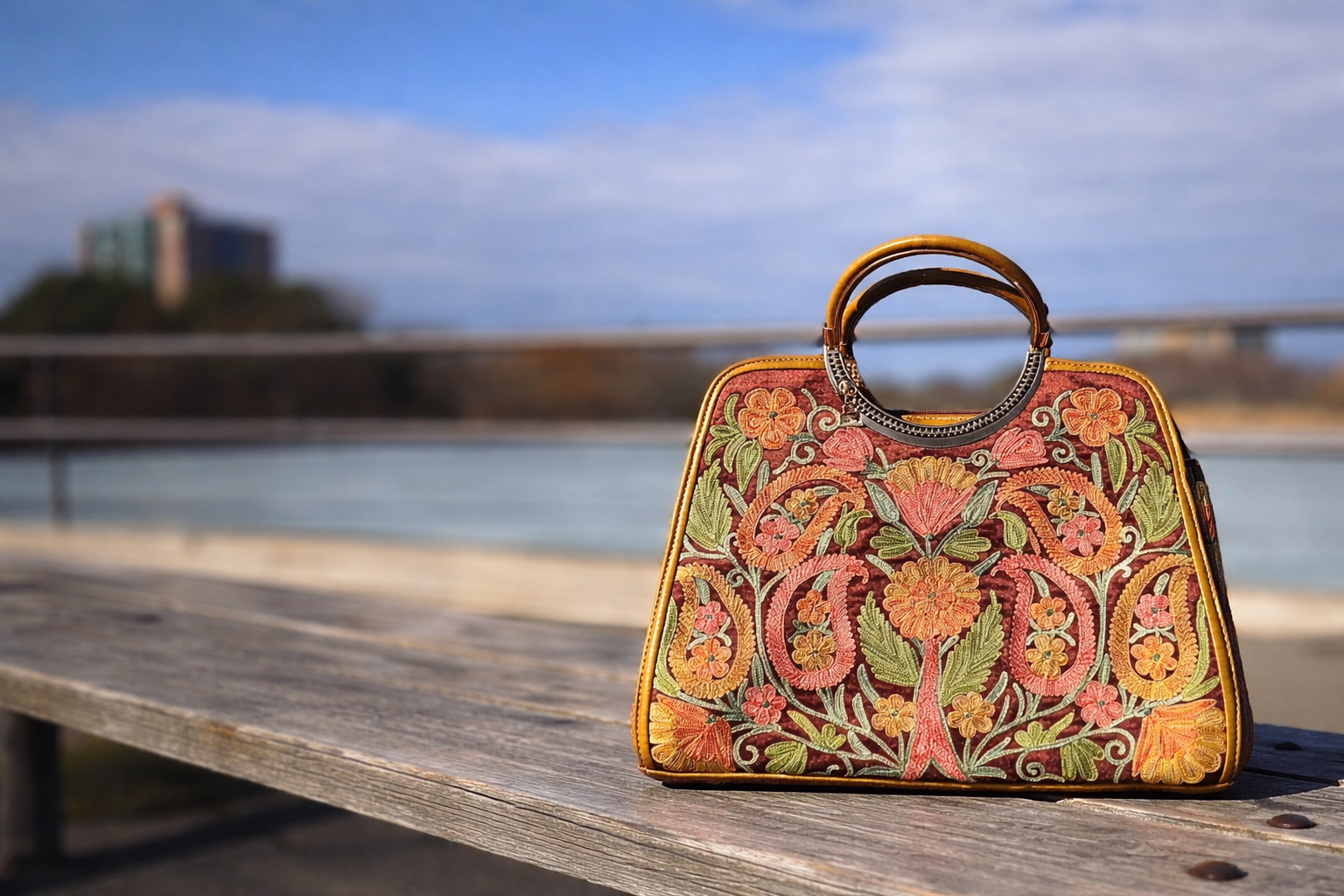 Decorative handbag with floral patterns on a wooden bench by a waterfront.